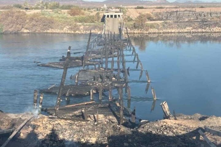 A burned-out wooden bridge, its skeletal remains jutting across a calm river, surrounded by scorched terrain and distant scrubland.