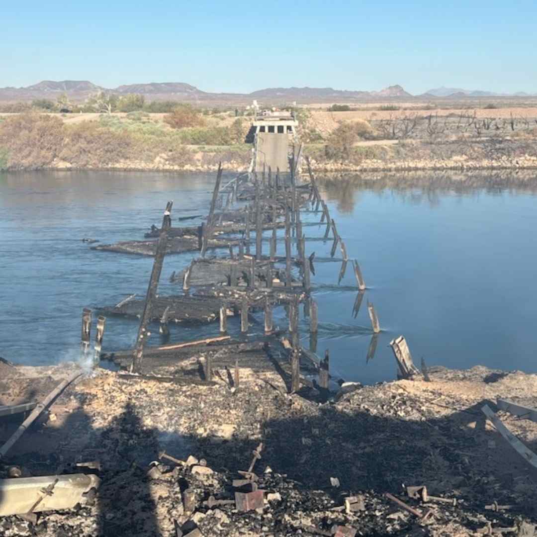 A burned-out wooden bridge, its skeletal remains jutting across a calm river, surrounded by scorched terrain and distant scrubland.