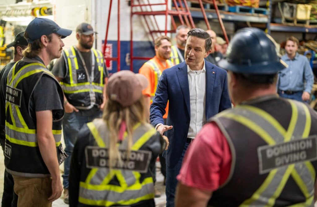 Pierre Poilievre talking with Dominion Diving workers in high-visibility vests inside a marine construction facility.
