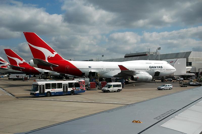 Multiple Qantas aircraft with red kangaroo tails parked at London Heathrow, ground vehicles on the tarmac, and an aircraft wing visible in the foreground.