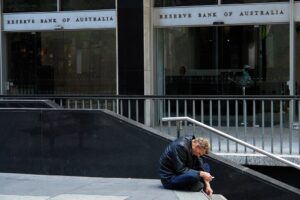 A man sitting on a low wall outside the Reserve Bank of Australia building, hunched forward with his head in his hands.