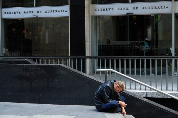 A man sitting on a low wall outside the Reserve Bank of Australia building, hunched forward with his head in his hands.