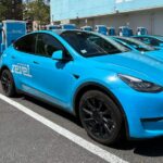 A lineup of blue electric Revel vehicles parked at a charging station, with city buildings in the background and the Revel logo visible on-site.
