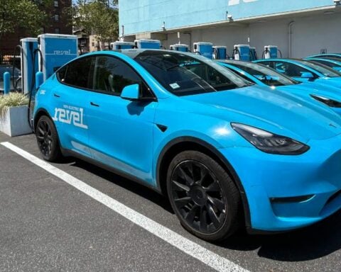 A lineup of blue electric Revel vehicles parked at a charging station, with city buildings in the background and the Revel logo visible on-site.