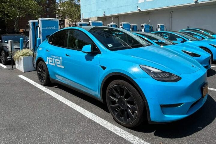 A lineup of blue electric Revel vehicles parked at a charging station, with city buildings in the background and the Revel logo visible on-site.
