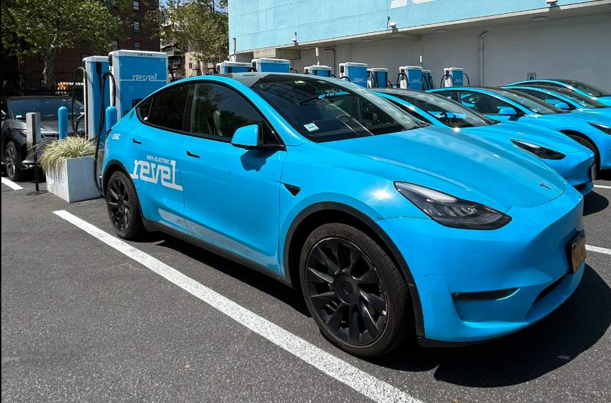 A lineup of blue electric Revel vehicles parked at a charging station, with city buildings in the background and the Revel logo visible on-site.