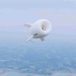 White torus‑shaped airborne wind turbine with stabilizing fins flying over mixed land and water landscape, branded “XUWEI,” credited to Xinhua News.