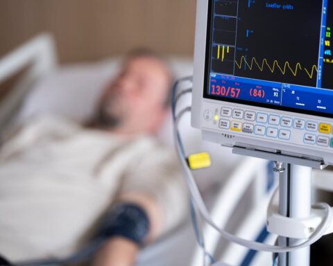 Hospital patient monitoring equipment showing vital signs with a heart rate monitor display and blurred patient in hospital bed in the background.