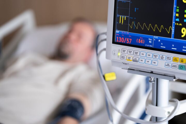 Hospital patient monitoring equipment showing vital signs with a heart rate monitor display and blurred patient in hospital bed in the background.