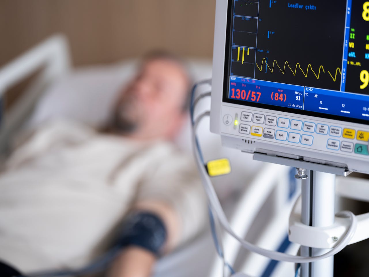 Hospital patient monitoring equipment showing vital signs with a heart rate monitor display and blurred patient in hospital bed in the background.