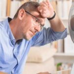 Person using a desk fan for cooling while seated with a laptop indoors, indicating heat-related HVAC strain