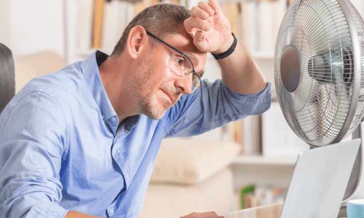 Person using a desk fan for cooling while seated with a laptop indoors, indicating heat-related HVAC strain