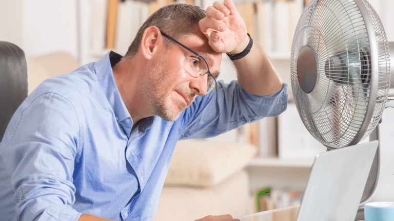 Person using a desk fan for cooling while seated with a laptop indoors, indicating heat-related HVAC strain