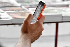 Close‑up of a person holding a smartphone with a red and black case, screen showing a grayscale image, in an outdoor setting with blurred stacks of newspapers or magazines in the background.