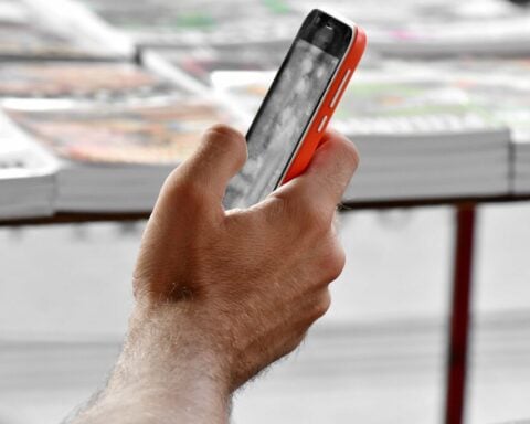 Close‑up of a person holding a smartphone with a red and black case, screen showing a grayscale image, in an outdoor setting with blurred stacks of newspapers or magazines in the background.