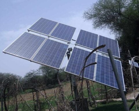 A ground-mounted solar panel array installed on a metal pole structure in a rural setting with trees in the background under a clear blue sky.