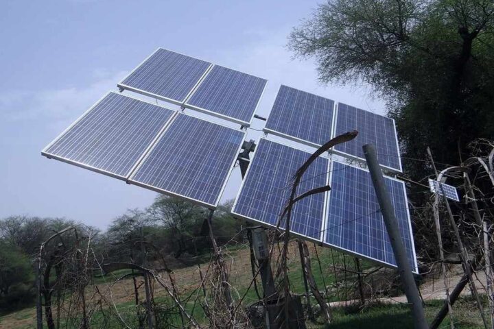 A ground-mounted solar panel array installed on a metal pole structure in a rural setting with trees in the background under a clear blue sky.