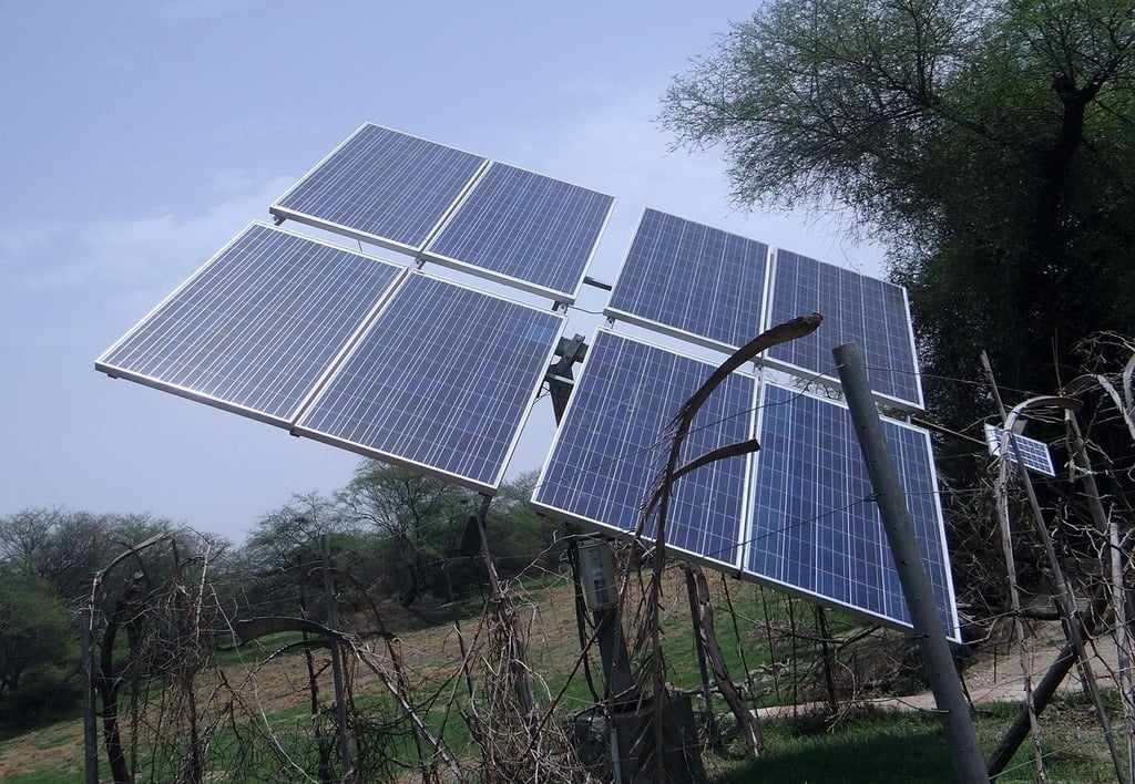 A ground-mounted solar panel array installed on a metal pole structure in a rural setting with trees in the background under a clear blue sky.