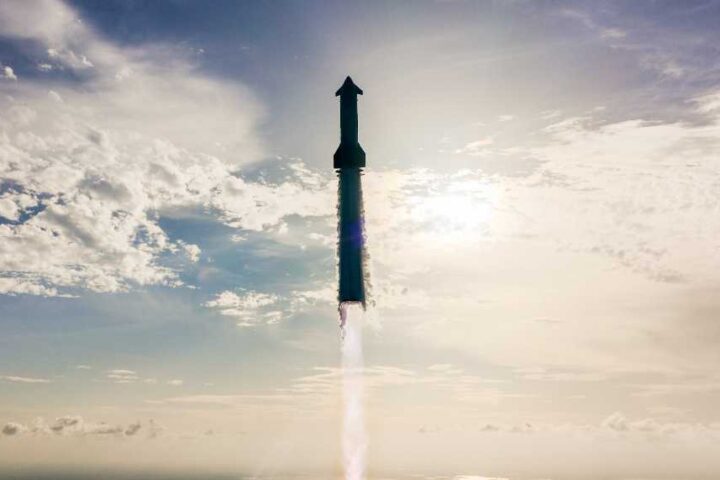 SpaceX Starship rocket ascending through clouds with engine firing during Flight 10 test, silhouetted against a bright blue sky with scattered white clouds.