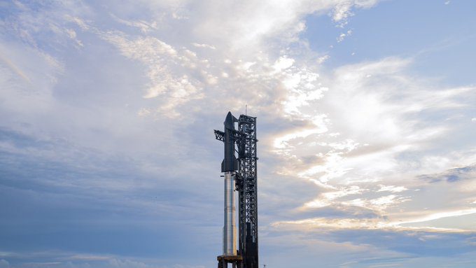 Fully stacked Starship and Super Heavy rocket at the launch pad under cloudy skies, with vapors surrounding the base as it awaits the Flight 10 test.