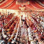 Rows of dozens of Indian brides and grooms in traditional attire seated side by side under a decorated canopy during a large-scale community mass marriage ceremony.