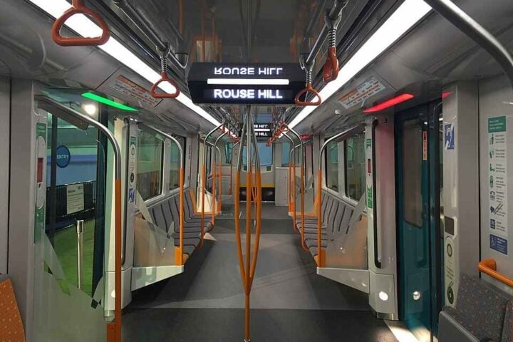 Interior of a Sydney Metro prototype train with overhead handrails, LED lighting, and bilingual digital displays showing “Rouse Hill.”