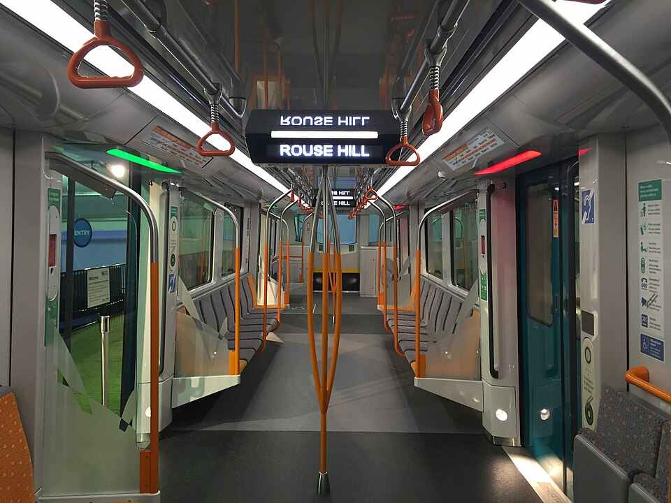 Interior of a Sydney Metro prototype train with overhead handrails, LED lighting, and bilingual digital displays showing “Rouse Hill.”