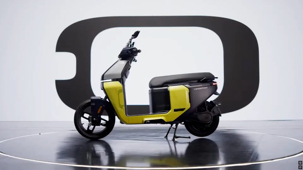 TVS Orbiter electric scooter in bright yellow and black color scheme displayed on a circular platform against a large black 'O' logo background, showing its distinctive boxy design, flat footboard, and 14-inch front wheel.