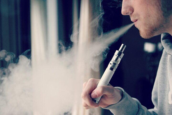 Person exhaling vapor cloud while holding an electronic cigarette against a dark background, illustrating the youth vaping concern linked to increased smoking risk.