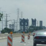 Power transmission lines and towers against a gray stormy sky in Chicago, with orange construction barriers visible in the foreground on a highway.