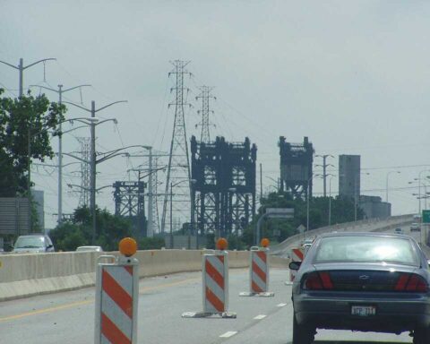 Power transmission lines and towers against a gray stormy sky in Chicago, with orange construction barriers visible in the foreground on a highway.