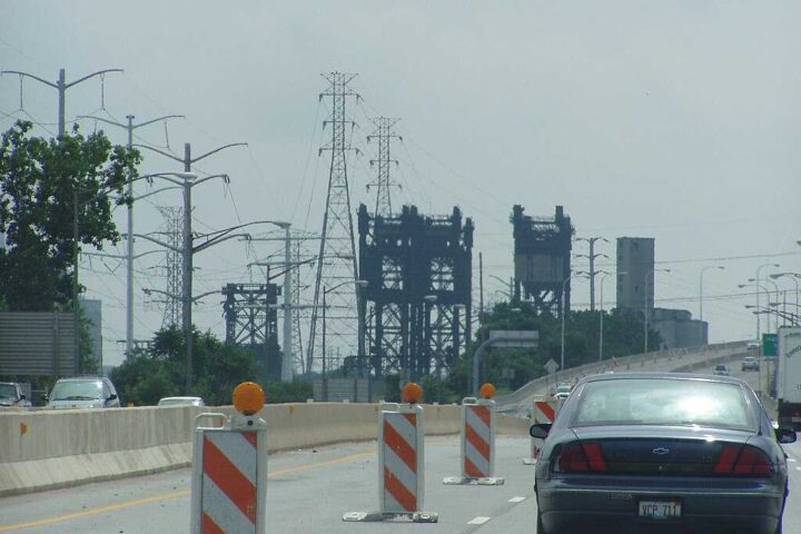Power transmission lines and towers against a gray stormy sky in Chicago, with orange construction barriers visible in the foreground on a highway.
