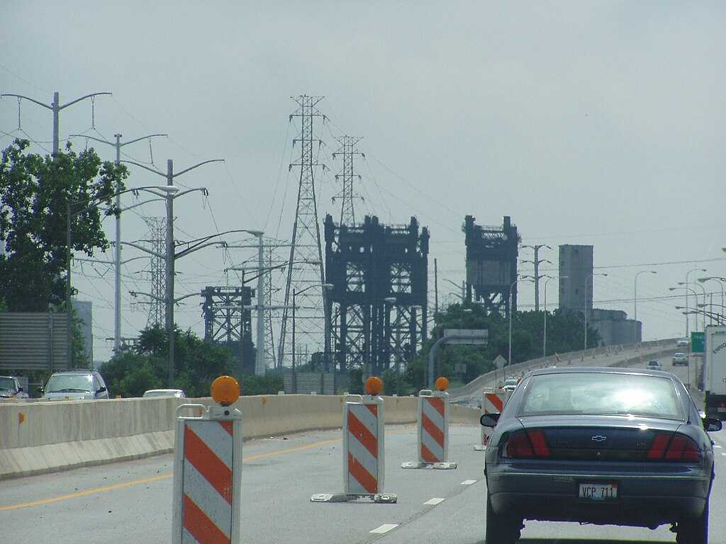 Power transmission lines and towers against a gray stormy sky in Chicago, with orange construction barriers visible in the foreground on a highway.
