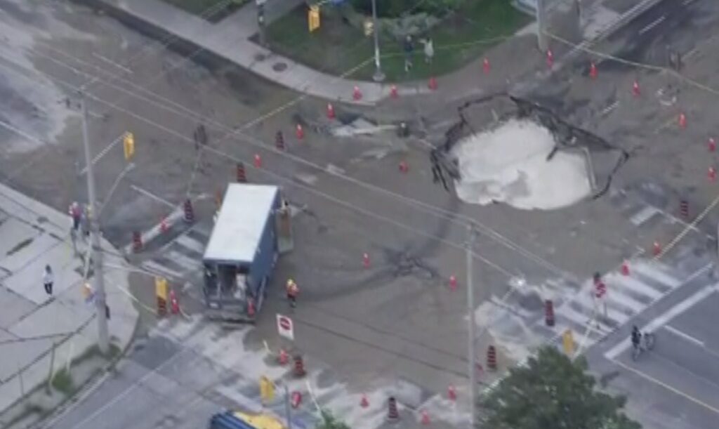 Aerial view of a large sinkhole at Coxwell Avenue and Cosburn Avenue in Toronto, surrounded by traffic cones and barriers, with repair crews on site and a TTC bus stopped nearby.