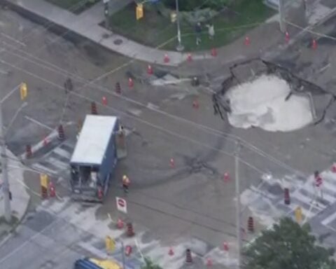 Aerial view of a large sinkhole at Coxwell Avenue and Cosburn Avenue in Toronto, surrounded by traffic cones and barriers, with repair crews on site and a TTC bus stopped nearby.