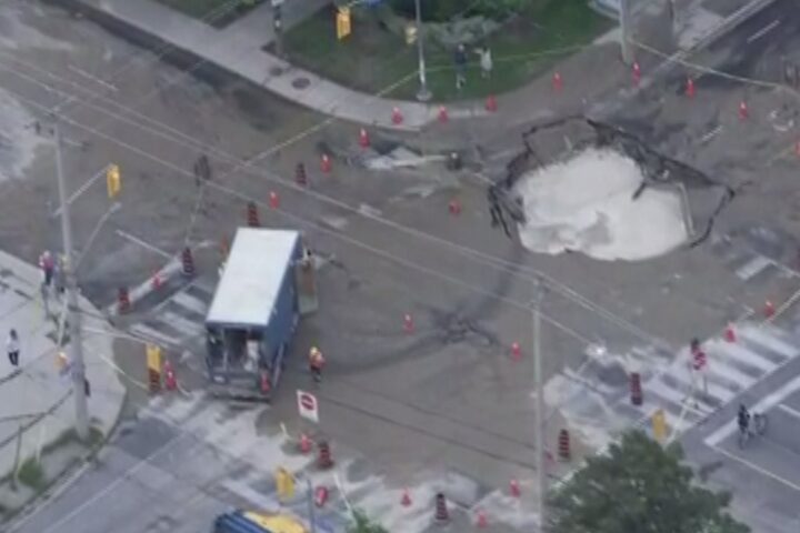 Aerial view of a large sinkhole at Coxwell Avenue and Cosburn Avenue in Toronto, surrounded by traffic cones and barriers, with repair crews on site and a TTC bus stopped nearby.