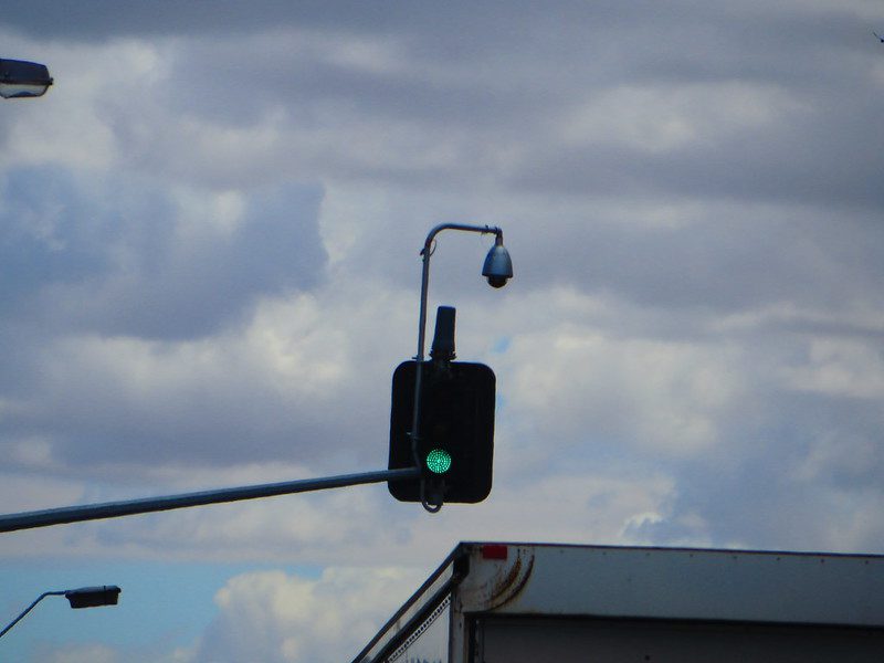 Overhead traffic-signal camera mounted above a green light at an Adelaide intersection, with cloudy sky background.