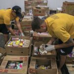 Two uniformed sailors in yellow shirts and gloves inspect and sort boxes of canned and packaged food inside a warehouse.