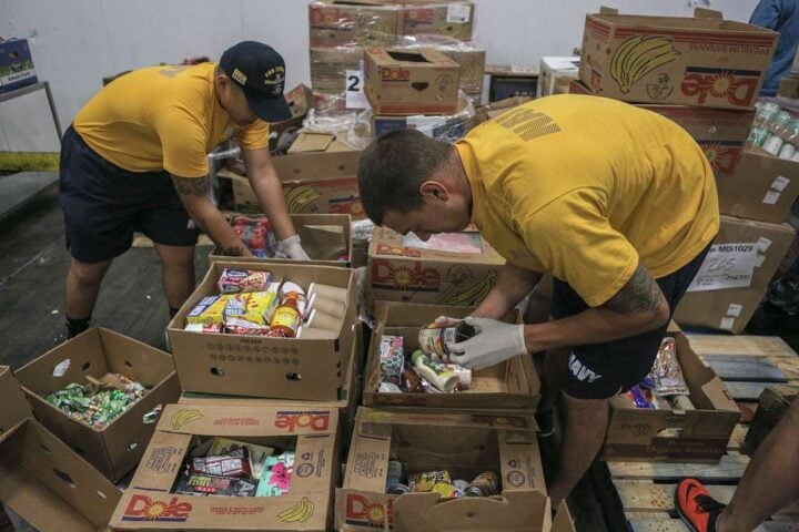 Two uniformed sailors in yellow shirts and gloves inspect and sort boxes of canned and packaged food inside a warehouse.