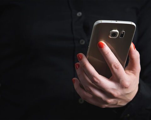 Person holding a smartphone in their hand with red nail polish, wearing a dark shirt against a black background.