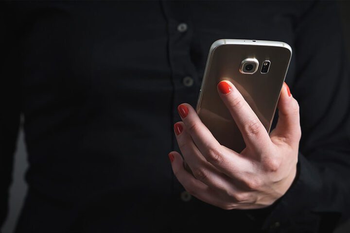 Person holding a smartphone in their hand with red nail polish, wearing a dark shirt against a black background.