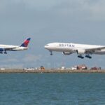 Delta Air Lines Boeing 757-200 and United Airlines Boeing 777-200 landing over water, runway equipment visible, mountains under cloudy sky.