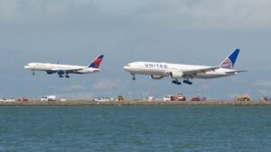 Delta Air Lines Boeing 757-200 and United Airlines Boeing 777-200 landing over water, runway equipment visible, mountains under cloudy sky.
