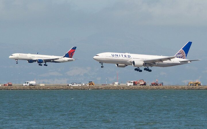 Delta Air Lines Boeing 757-200 and United Airlines Boeing 777-200 landing over water, runway equipment visible, mountains under cloudy sky.