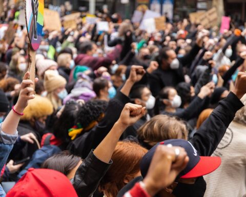 Crowd of protesters with raised fists in solidarity at a demonstration in Melbourne, Australia. Many wear face masks and hold signs, with diverse participants showing unity against discrimination.