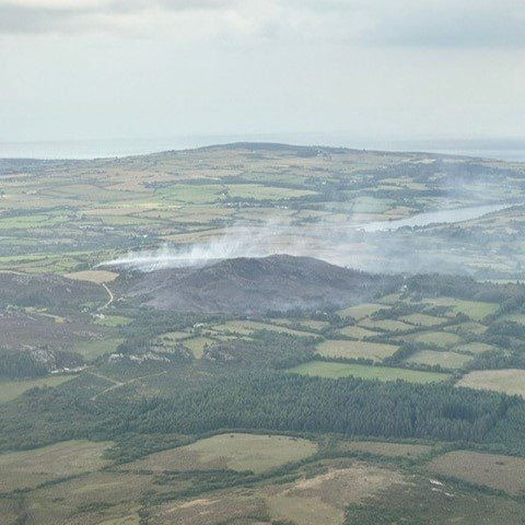 Aerial view of smoke rising from hills and countryside after a wildfire in North Wales, with fields and forests surrounding the affected area.