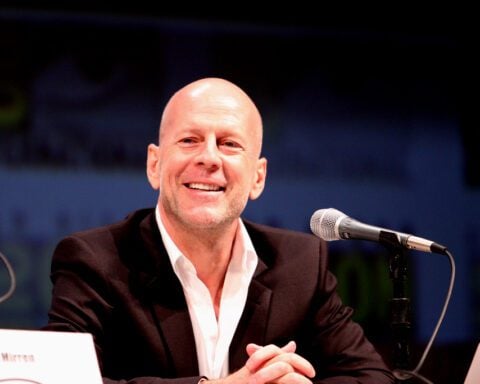 Bruce Willis smiling while seated at a panel during the 2010 San Diego Comic Con, wearing a dark suit with an open-collar white shirt.