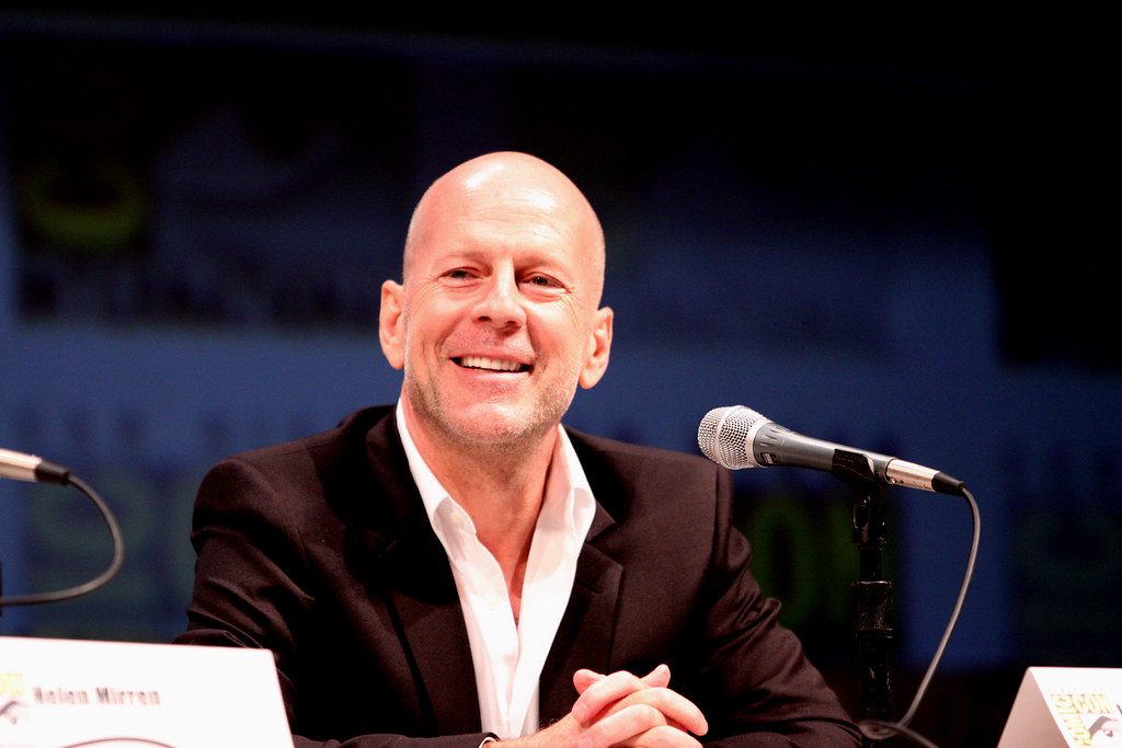 Bruce Willis smiling while seated at a panel during the 2010 San Diego Comic Con, wearing a dark suit with an open-collar white shirt.