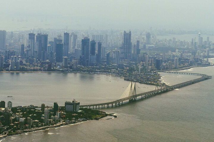 Mumbai skyline featuring high-rise buildings and the Bandra-Worli Sea Link bridge under a hazy sky.