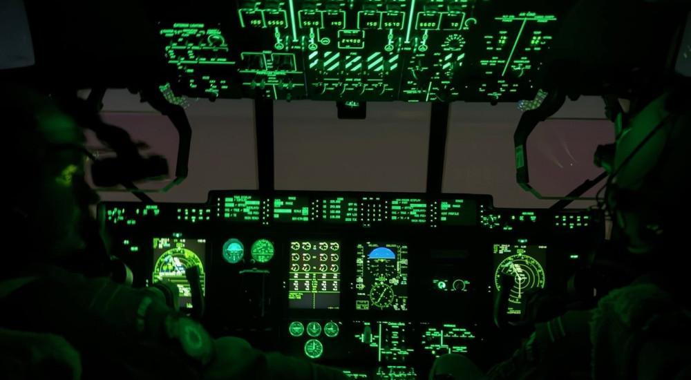 Two RNZAF pilots wearing helmets and night-vision goggles sit in a dimly lit Hercules cockpit with green instrument displays glowing.